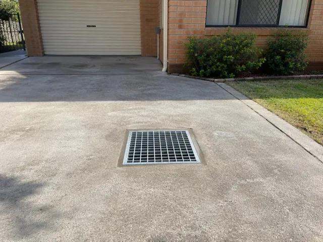 Dry concrete driveway with a square outdoor drain grate beside a brick house in Carramar